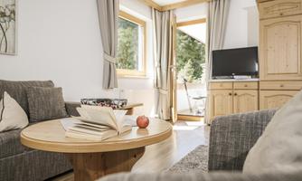 A cozy living room with a round wooden table and a reading nook. Large windows provide plenty of natural light and a view of the outside.