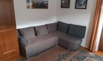 A cozy living room with a gray corner sofa and a carpet. Some pictures are hanging on the wall, and there is a wooden cupboard.