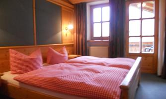 A cozy bedroom with a double bed and wooden paneling. The bed linen is red checkered, and there are large windows with curtains.