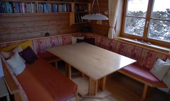 A cozy room with a wooden table and a U-shaped bench. The walls are made of wood and there is a view of a snowy landscape through the window.
