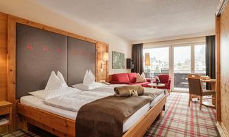 A cozy hotel room with a large bed and rustic wooden design. There is a balcony with a view and a small area with a sofa and a table.