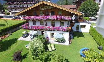 A charming building with a balcony full of flowers. The garden is green and inviting, surrounded by mountains.