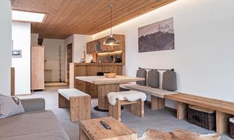 A modern, cozy living room with wooden interiors. There is a dining area with a bench and an open kitchen area.