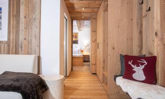 A cozy wooden hallway with a comfortable seating area and a decorative cushion. In the background, a bright room is visible, which looks inviting.
