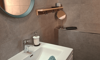 A modern bathroom with gray tiles and a round mirror. A turquoise towel lies on the sink and there is a wooden towel rack. Behind the door is the shower.