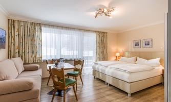 A bright room with a large bed, a sofa, and a table. Plenty of daylight through the windows with curtains.