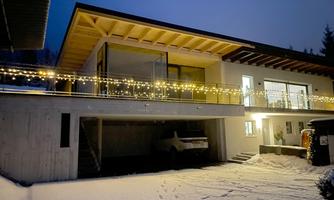 A modern house in a snowy setting, adorned with lights. The sky is dark blue and it is evening.