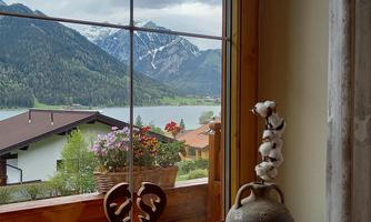 A view through a window of a mountain landscape with snow-capped peaks and a clear lake. In the foreground, there are flower pots and a decorative vase.