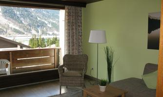 A bright living room with a cozy armchair and a table. Large windows offer a view of the mountains and a balcony.