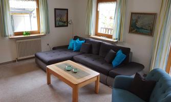 A cozy living room with a large couch and colorful cushions. A wooden table is in the center and windows let in natural light.