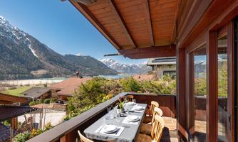 A terrace with a dining table and chairs, surrounded by mountains and a clear blue sky. In the background, snow-capped peaks and a calm lake can be seen.