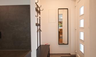 A modern hallway with light gray walls and a large mirror. There are coat hooks on the wall and a bright entrance door with windows.