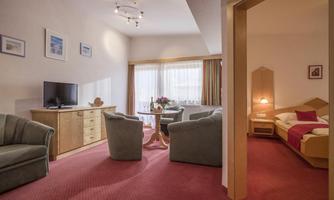 A cozy hotel room with a living area and a bedroom. There is a television, a table, and comfortable armchairs in a bright atmosphere.
