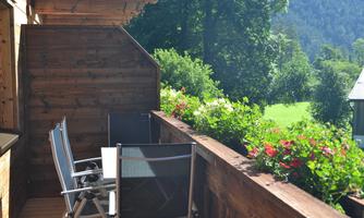 A cozy balcony with wooden cladding and blooming flowers. In the background, a green meadow and trees can be seen.
