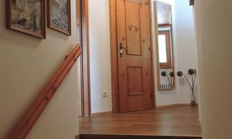 A bright hallway with wooden stairs and a door at the end. Pictures hang on the wall, and a window lets in natural light.