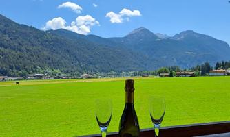 A picturesque view of green meadows and mountains under a clear blue sky. Two glasses and a bottle of champagne are in the foreground.