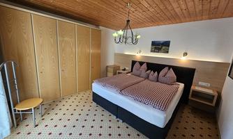 A cozy bedroom with a large bed and a wooden ceiling. In the background, a wardrobe and a stool can be seen.