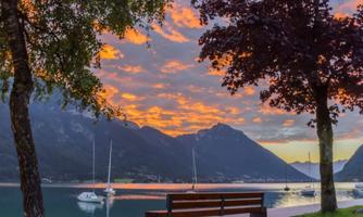 A picturesque lake with a bench in the foreground and gentle mountains in the background. The sky is colored with orange and purple clouds.