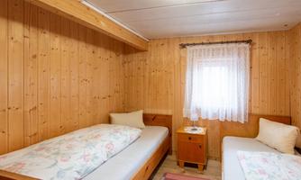 A simple bedroom with two single beds and wood paneling. The window is adorned with a white curtain and a nightstand stands between the beds.
