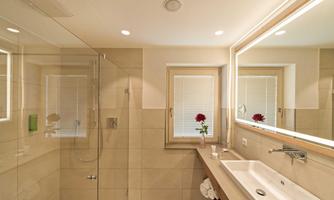 A modern bathroom with a glass shower and bright tiles. The sink area is simple and a small bouquet of flowers is on the windowsill.