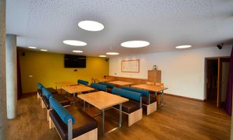 A modern room with wooden tables and blue benches. The walls are painted in soft colors and there are round ceiling lights.