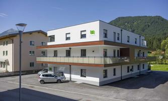 A modern apartment building with a bright facade and large windows. In the background, green hills are visible.