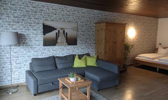 A modern living room with a gray sofa, a wooden coffee table, and a floor lamp. The walls are decorated with patterned wallpaper and a bed is visible in the background.