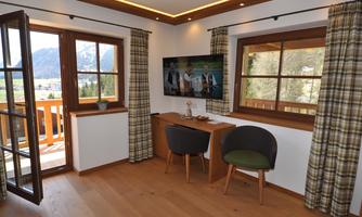 A cozy room with wooden decor and large windows. A desk and a television are also visible, overlooking a mountainous landscape.