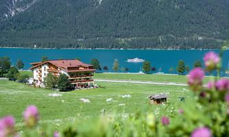 A picturesque landscape with a hotel by the water and a clear, turquoise lake. In the background, green meadows and mountains can be seen.
