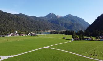 A green meadow surrounded by high mountains and a clear blue sky. In the background, houses and a pond are visible.