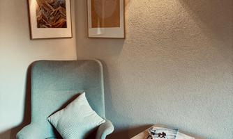 A cozy armchair is placed in a quiet corner, complemented by a small table. Two pictures hang on the wall, while soft wall lighting brightens the room.