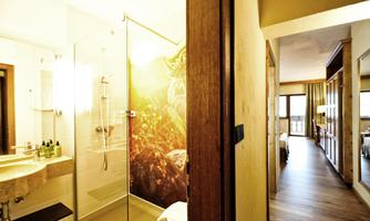 A modern bathroom with a glass shower and sink. In the background, there is a bright hallway with wooden flooring and access to additional rooms.