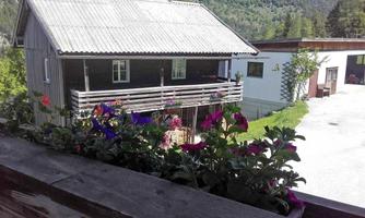 A traditional wooden house with a balcony and colorful flowers in the foreground. In the background, green mountains are visible.