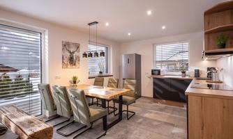 A modern kitchen with a large dining table and comfortable chairs. Large windows provide plenty of daylight and a view outside.