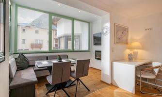 A bright living room with large windows and modern furniture. There is a dining area, a desk, and plenty of space for relaxation.