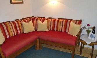 A cozy corner sofa with red-striped cushions is set in a bright room. On a table next to the sofa, there are a bottle and floral arrangements.