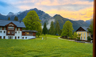 A beautiful view from the window of a green meadow with trees and majestic mountains in the background. The sky is illuminated in warm colors during sunset.