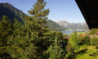 A picturesque landscape with green trees and a view of a lake. In the background, majestic mountains are visible under a clear blue sky.