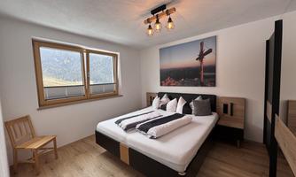 A modern bedroom with a double bed and decorative pillows. Large windows provide a view of the mountains.