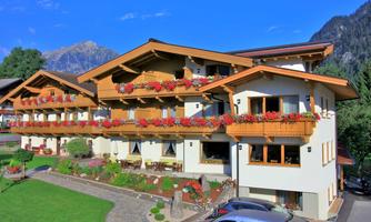 A beautiful alpine building with wooden cladding and balconies adorned with colorful flowers. In the background, green meadows and mountains can be seen.