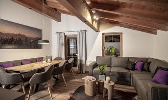 A modern living room with a wooden table and comfortable gray chairs. The room has a cozy atmosphere with a wood-paneled ceiling and decoration in purple colors.