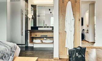 A modern bathroom with wooden shelves and a sink. A white bathrobe hangs on a hook next to a fabric bag.