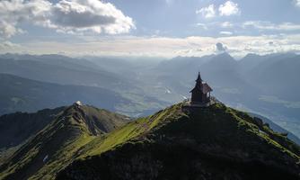 A small chapel on a mountain peak surrounded by impressive landscape. In the background, the surrounding mountains and the sky are visible.