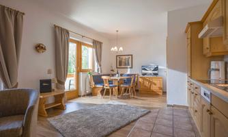 A cozy living room with wooden furniture and a dining table. Large windows provide plenty of daylight.