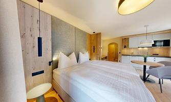 A modernly designed hotel room with a large bed and wooden paneling. In the background, there is a small kitchenette and a dining area.