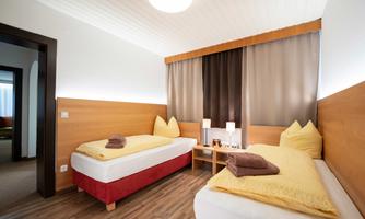 A cozy room with two single beds, wooden paneling, and modern lamps. The windows are decorated with light curtains and the floor is made of wood.