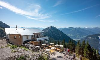 A mountain inn with sun umbrellas and outdoor seating. The view shows majestic mountains and a clear blue sky.