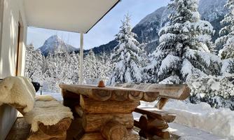 A wooden chair and a table are on a terrace overlooking a snowy landscape. Tall, snow-covered trees and mountains form the background.