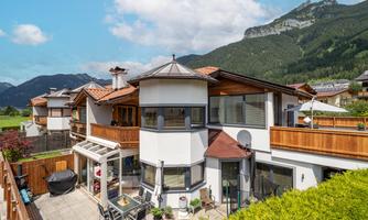 A modern house in the mountains with a beautiful garden and a terrace. The landscape is green and the mountains are visible in the background.
