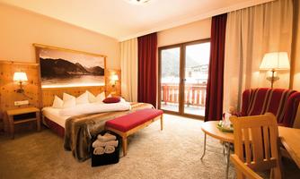 A cozy hotel room with a large bed and wooden details. The room has large windows with a view outside and stylish furniture.